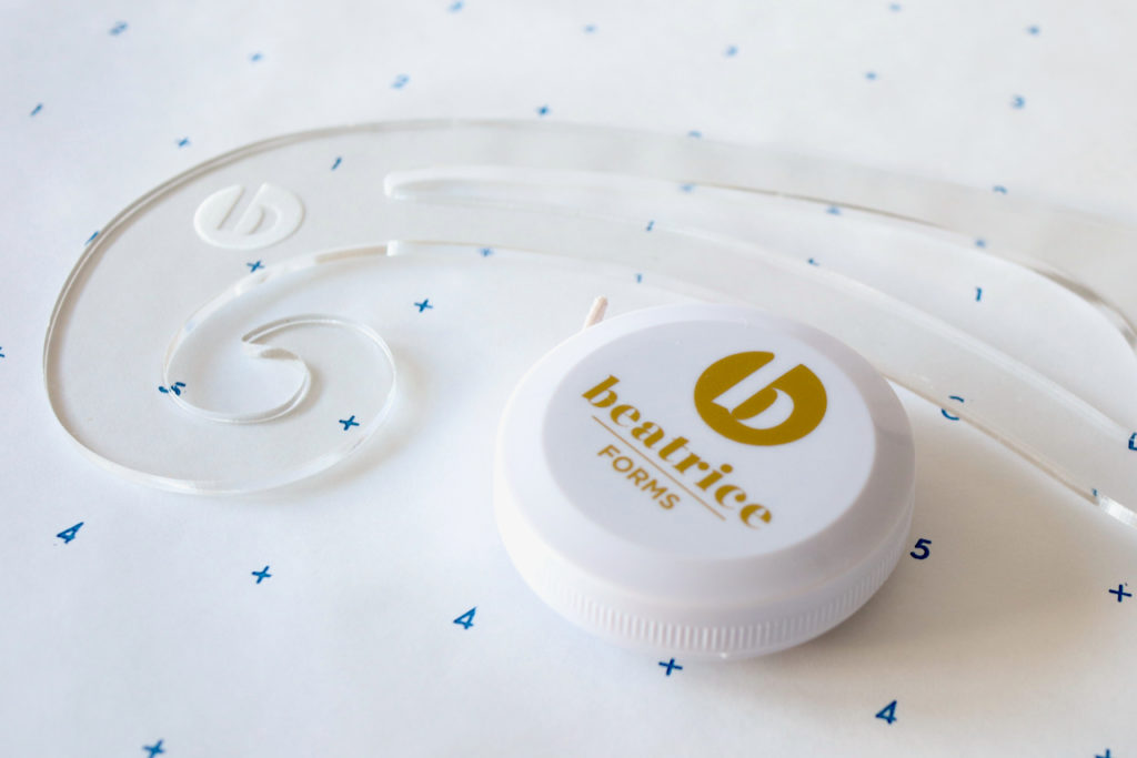 A french curve ruler and a retractable measuring tape with the Beatrice Forms logo sitting on printed pattern paper